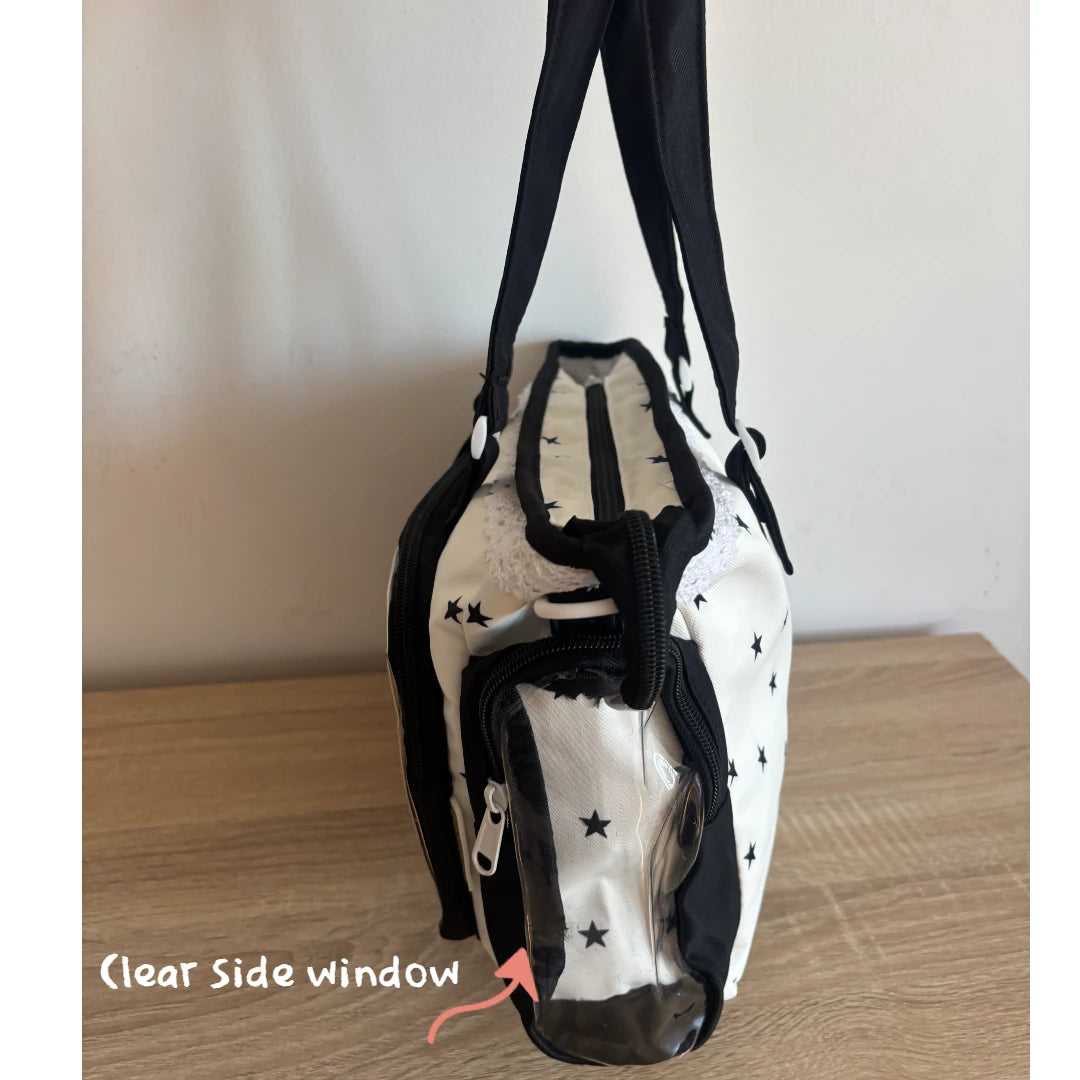 A white and black Ita shoulder bag with star patterns, featuring a clear side window labeled 'Clear Side window' with a pink arrow, shown standing on a wooden surface against a plain wall.