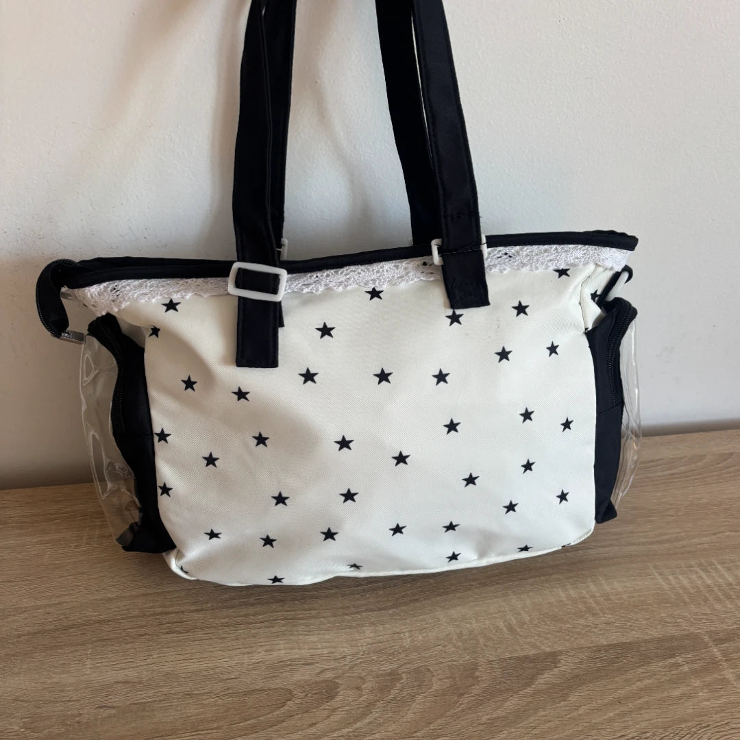 An ita bag with a white canvas body covered in small black stars, black fabric handles, a black top trim, and clear plastic side panels, shown resting on a wooden surface against a plain wall