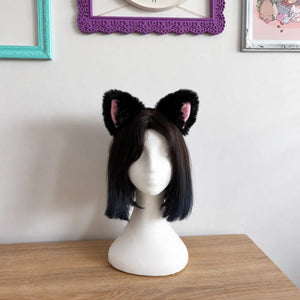 A black faux-fur cat ear headband worn with a short black bob wig on a white mannequin head, displayed on a wooden table against a white wall with framed pictures.