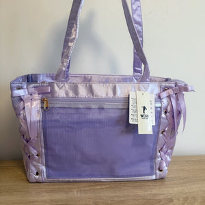 A lavender purple ita bag with a mesh front pocket, shiny satin handles, and lace-up side details, shown with a hangtag on a wooden surface against a plain wall.