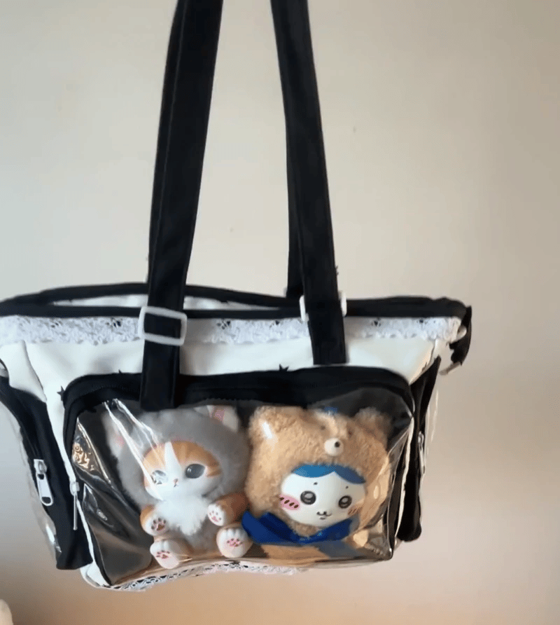 A clear window Ita bag with black handles and trim, containing two plush toys, photographed against a plain beige wall.