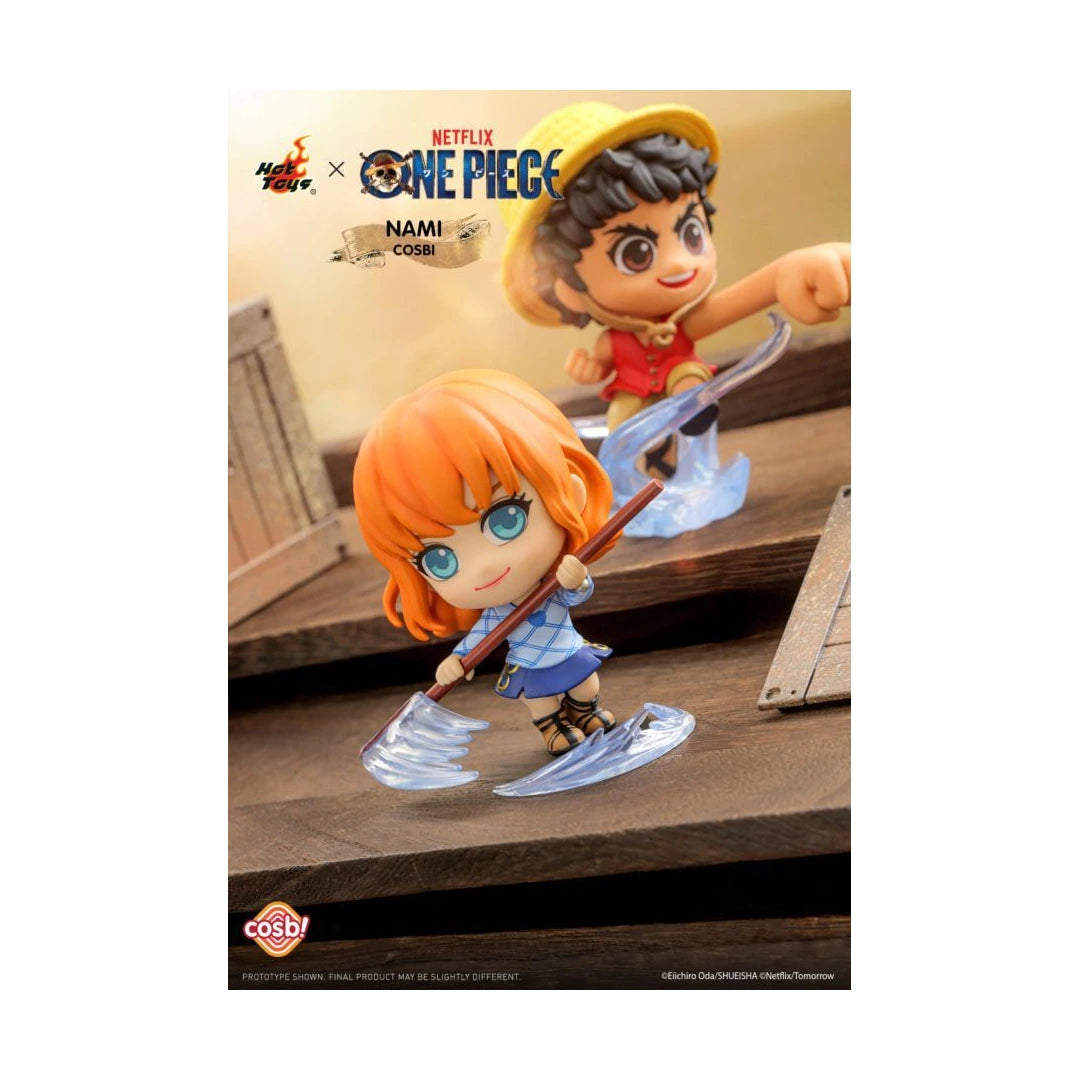 A Hot Toys x One Piece Nami collectible figure set shown on a printed backdrop card. The front figure has orange hair, blue clothing, and holds a shovel with clear ice effects, while a second figure in a straw hat appears behind her.