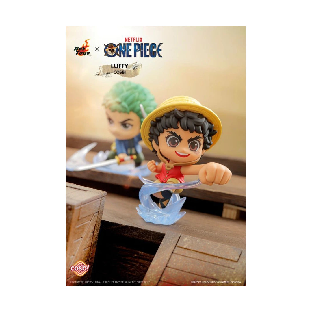 Hot Toys x One Piece Luffy Cosplay collectible figure shown in a themed diorama setting with a wooden crate and a blurred background figure. The figure wears a yellow straw hat and red outfit with a blue wave base.