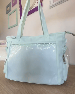 A light mint green ita bag with a glossy clear plastic front pocket, shown sitting on a wooden floor against a beige wall with framed pictures.