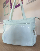 A light mint green diaper bag with a glossy clear plastic front pocket, shown sitting on a wooden floor against a beige wall with framed pictures.