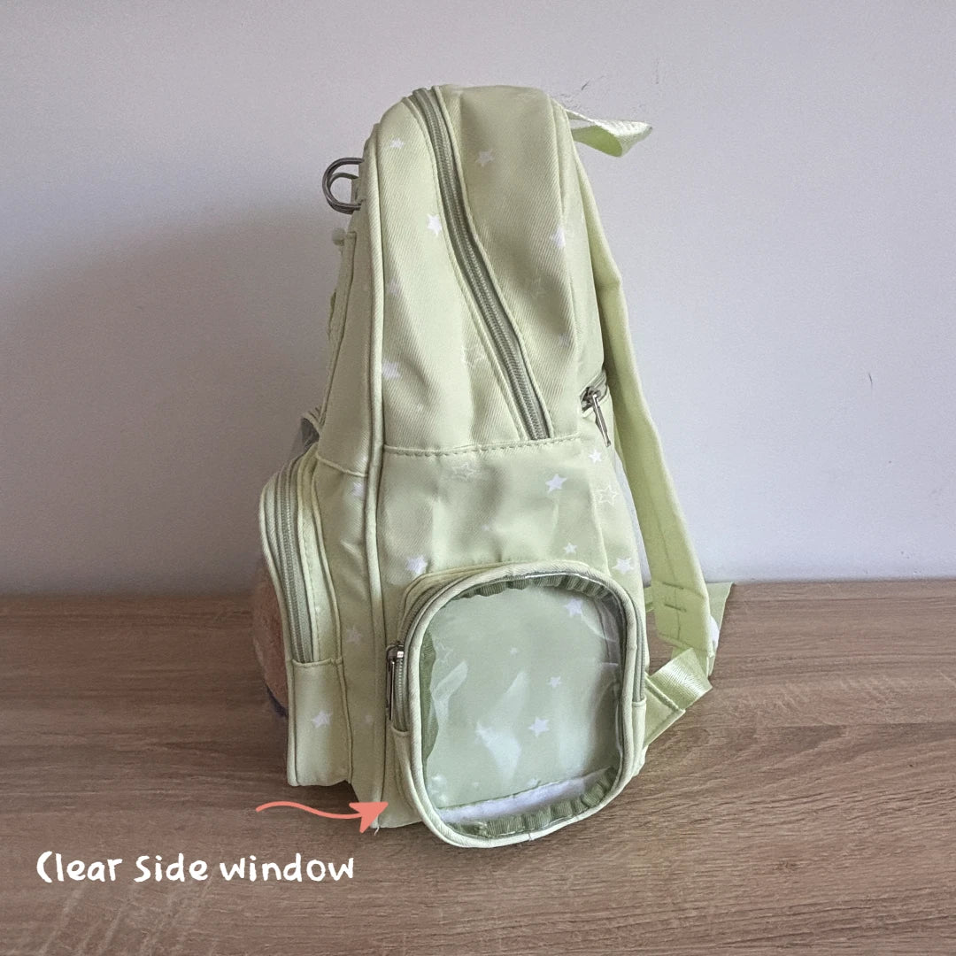 A light green Ita backpack with a star pattern, shown standing on a wooden surface against a plain wall. The image highlights a clear side window with an orange arrow and text overlay reading 'Clear Side window'.