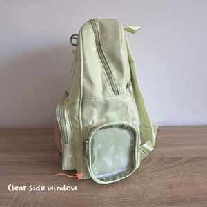A light green Ita backpack with a star pattern, shown standing on a wooden surface against a plain wall. The image highlights a clear side window with an orange arrow and text overlay reading 'Clear Side window'.