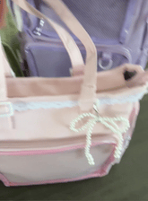 A close-up of a light pink Ita bag with a white pearl bow charm, white lace trim, and pink zipper accents, with a purple bag in the blurred background.