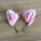 Pink and white faux fur cat ears headband on a light brown wood grain background.