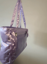 A lavender satin pin bag with side lacing and silver-tone hardware, shown hanging against a plain off-white wall.