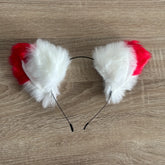 A pair of cat ears headband featuring fluffy white faux fur and bright red fabric tops, attached to a thin black wire band, shown on a light brown wood grain surface.