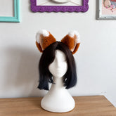 A short black bob wig with fluffy brown and white fox ears on a white mannequin head, displayed on a wooden table against a white wall with framed pictures.
