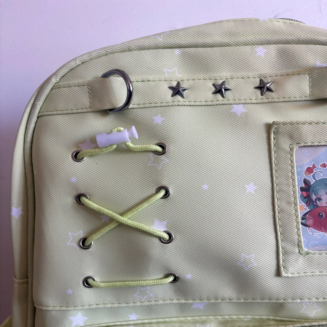 Close-up of a light green canvas backpack with white star patterns, featuring decorative metal eyelets with light green laces, silver star studs, and a cartoon patch on the side pocket.