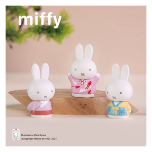 Three miffy figurines in colorful outfits on a wooden stand with a beige background.