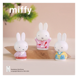 Three miffy figurines in colorful outfits on a wooden stand with a beige background.