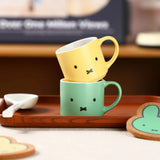 Two colorful mugs with Miffy the bunny face designs stacked on a wooden tray.