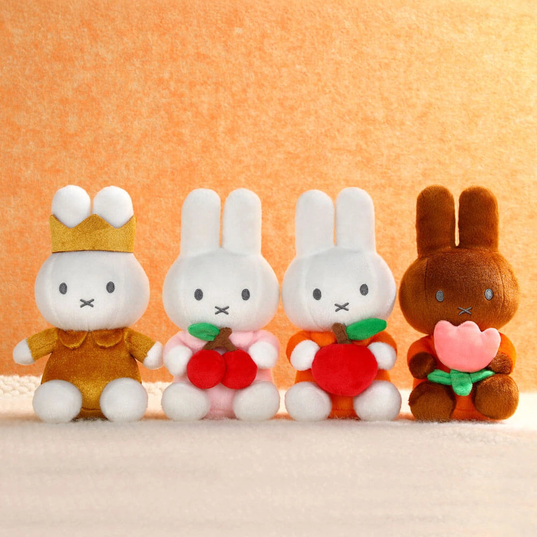Four Miffy plush toy bunnies with different colored bodies and accessories on a beige surface.