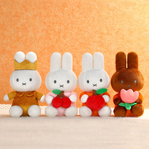 Four Miffy plush toy bunnies with different colored bodies and accessories on a beige surface.