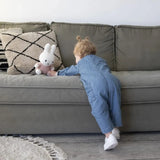 Child in blue outfit reaching for a pink plush toy on a gray sofa.