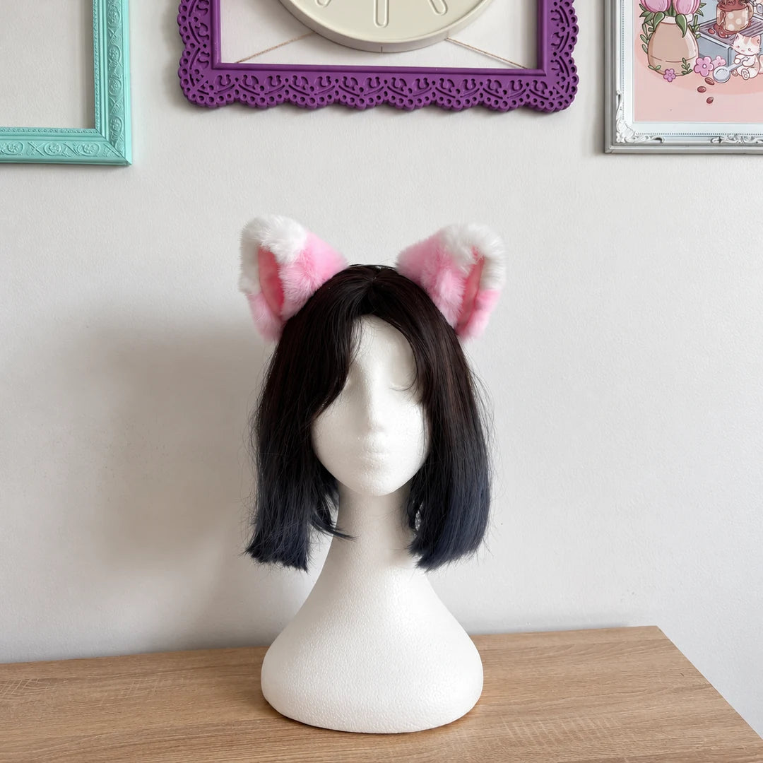 A short black bob wig with pink and white fluffy cat ears on a white mannequin head, displayed on a wooden table against a white wall with framed pictures.