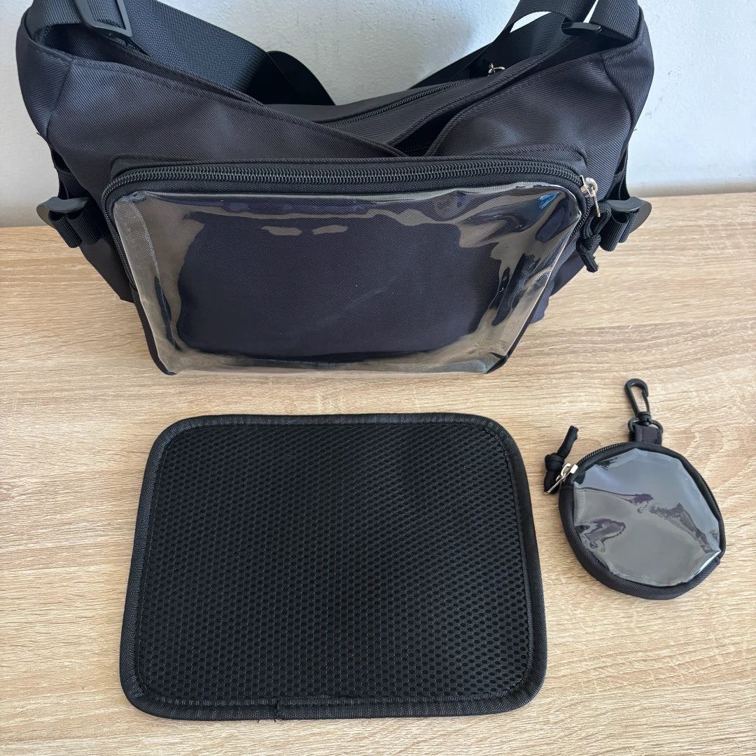 Black ita crossbody bag with a clear front pocket, shown on a light wood tabletop with a matching black mesh pouch and a small round clear zip pouch.