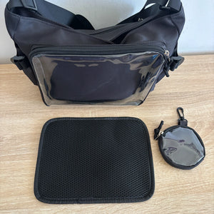 Black ita crossbody bag with a clear front pocket, shown on a light wood tabletop with a matching black mesh pouch and a small round clear zip pouch.