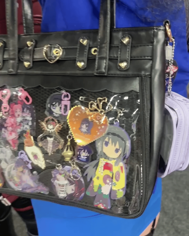 A black faux-leather ita bag with a clear front mesh pocket filled with colorful anime character keychains and charms, worn by a person in a blue outfit.