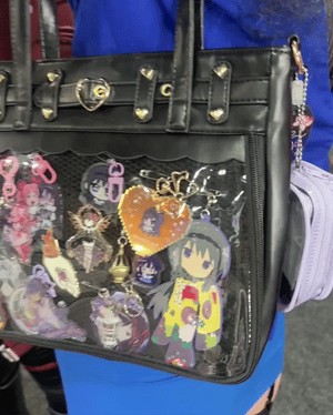 A black faux-leather ita bag with a clear front mesh pocket filled with colorful anime character keychains and charms, worn by a person in a blue outfit.