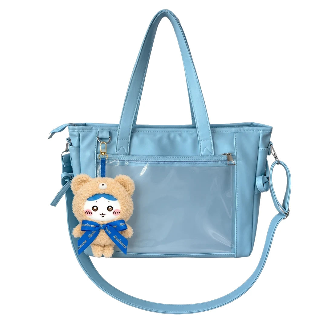 A light blue ita bag with a transparent front pocket, dual top handles, a detachable shoulder strap, and a plush teddy bear keychain with a blue bow hanging from the side, shown on a plain white background.