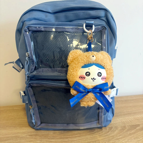 A blue mesh ita backpack with a cute plush character keychain hanging from the top zipper. The keychain features a brown teddy bear face with a white muzzle, pink cheeks, and a blue ribbon bow.