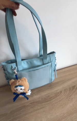 A light blue Ita shoulder bag with straps, held by a hand at the top left, resting on a light wood surface against a white wall. A small plush keychain with a blue ribbon is attached to the side.
