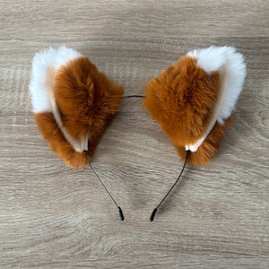 A pair of fluffy faux-fur animal ears, styled like a fox or rabbit, in brown and white, attached to thin black headbands, displayed on a light wood floor.