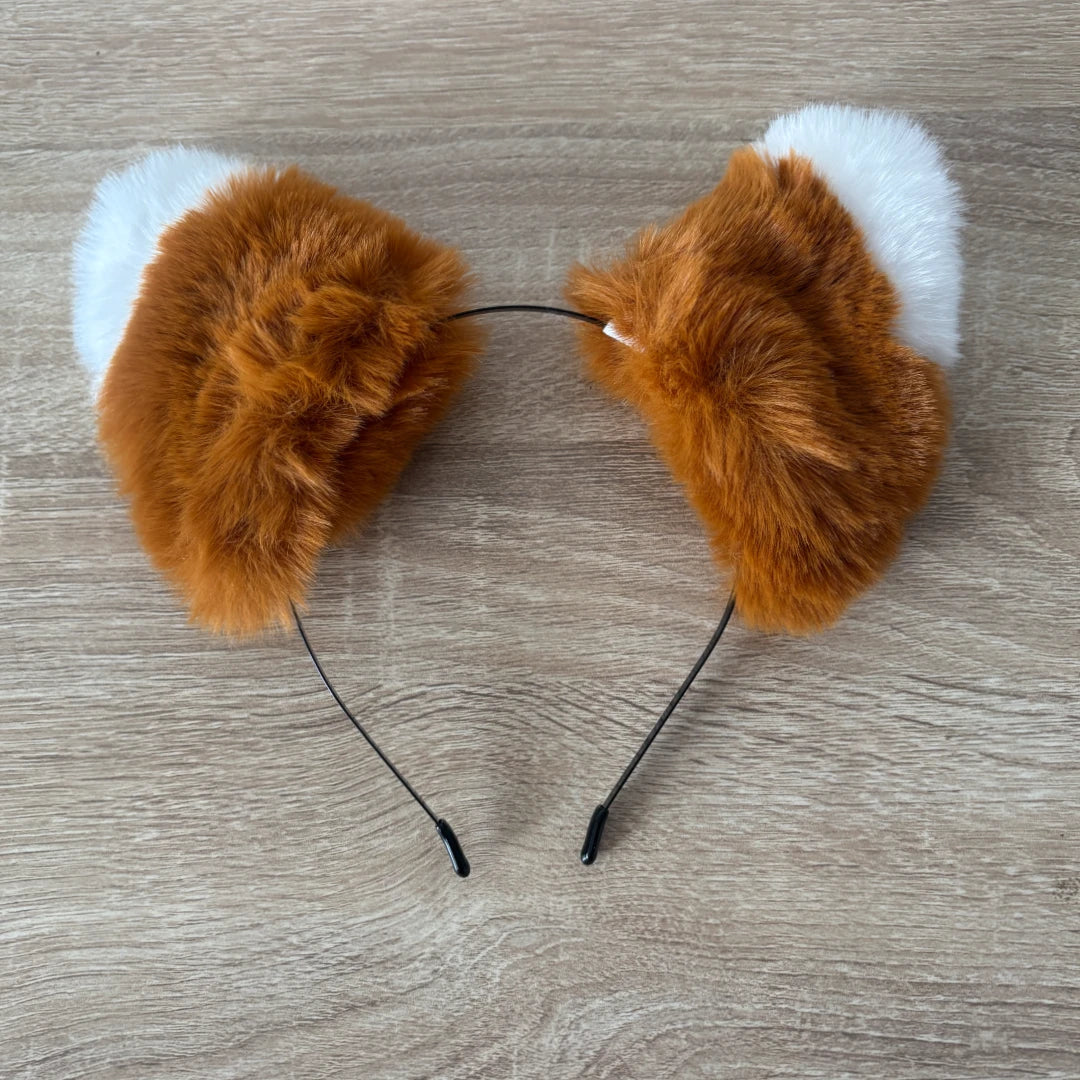 A pair of fluffy faux-fur headband accessories shaped like fox ears, with orange-brown fur on the front and white fur on the back, attached to a thin black elastic band, shown on a light wood surface.