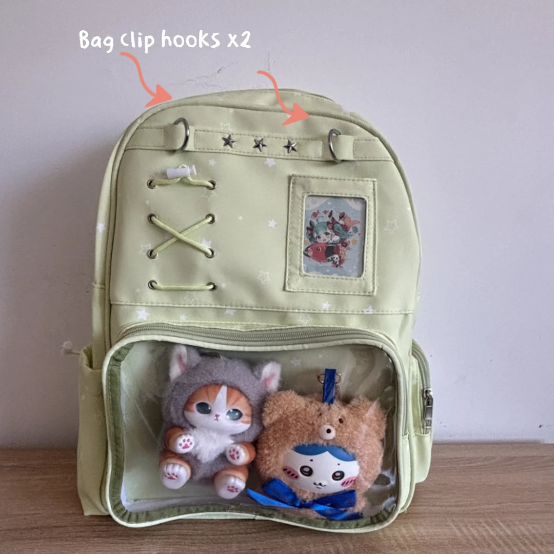 A pale yellow-green Ita backpack with a clear plastic front pocket containing two plush toys, shown against a plain wall with text reading 'Bag clip hooks x2' and arrows pointing to metal clips on the top.