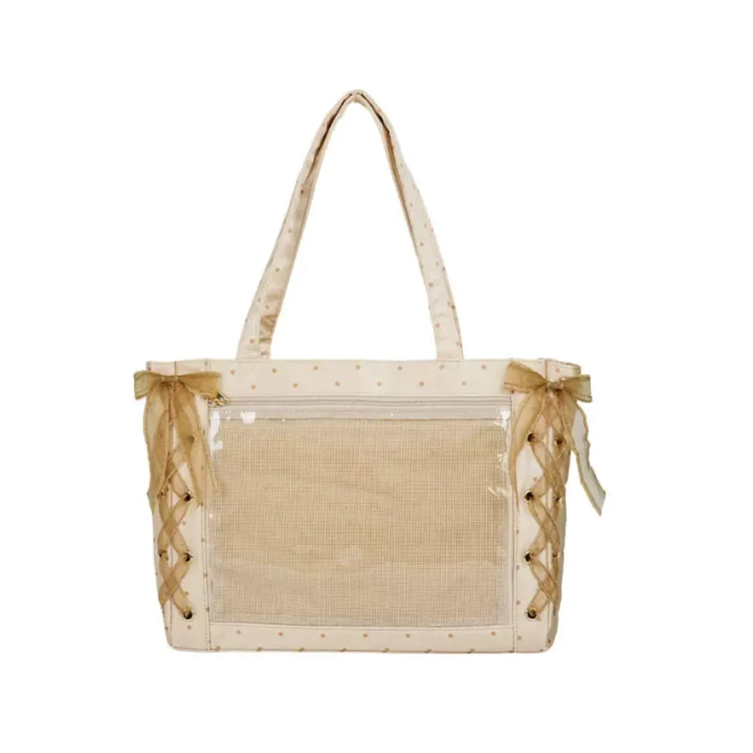 A cream-colored itabag with a clear front pocket, decorated with small gold polka dots, sheer ribbon handles, and lace-up side details.