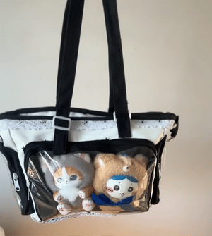 A white Ita bag with a black starred pattern with black handles and trim, containing two plush toys, photographed against a plain beige wall.