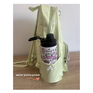 A pale yellow-green Ita backpack with a side water bottle pocket holding a white insulated bottle with a black lid and pink cartoon print. The photo is taken on a wooden surface against a plain wall, with the text 'Water bottle pocket' and an arrow pointing to the pocket.