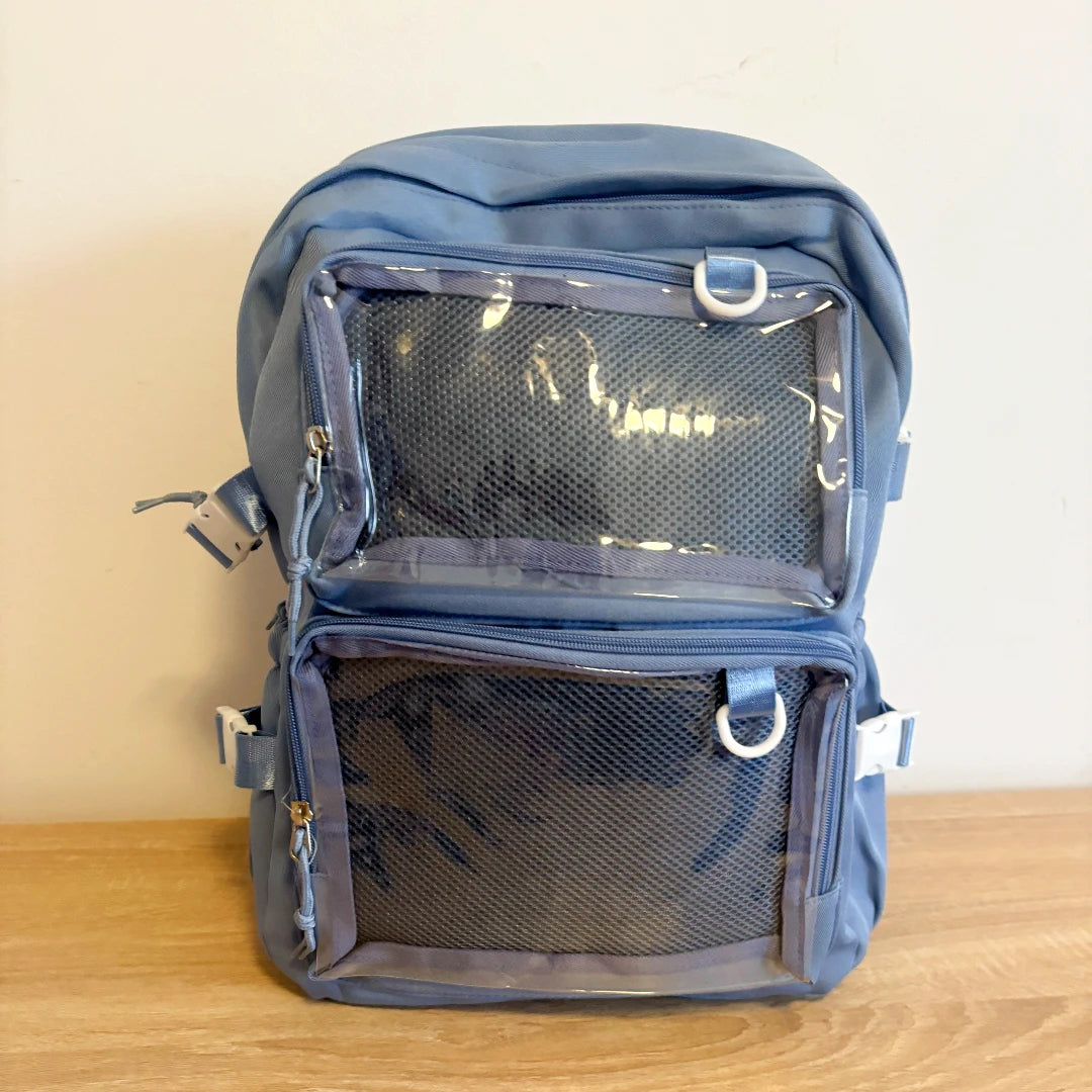 A blue Ita backpack with two front mesh document pockets, shown standing on a wooden surface against a plain beige wall.