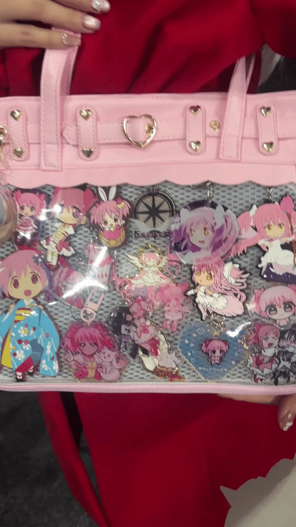 A pink ita bag with a clear mesh front panel covered in colorful anime character charms and pins, held by a person wearing a red dress.