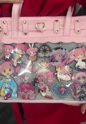 A pink ita bag with a clear acrylic front panel filled with various anime character charms and keychains, held by a person wearing a red top.