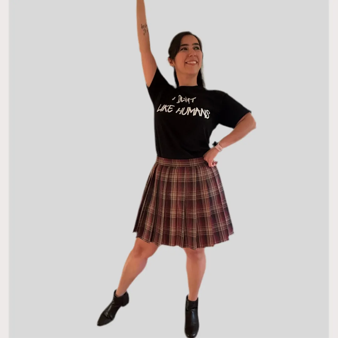 Person wearing a black t-shirt with text and a brown plaid skirt on a plain background