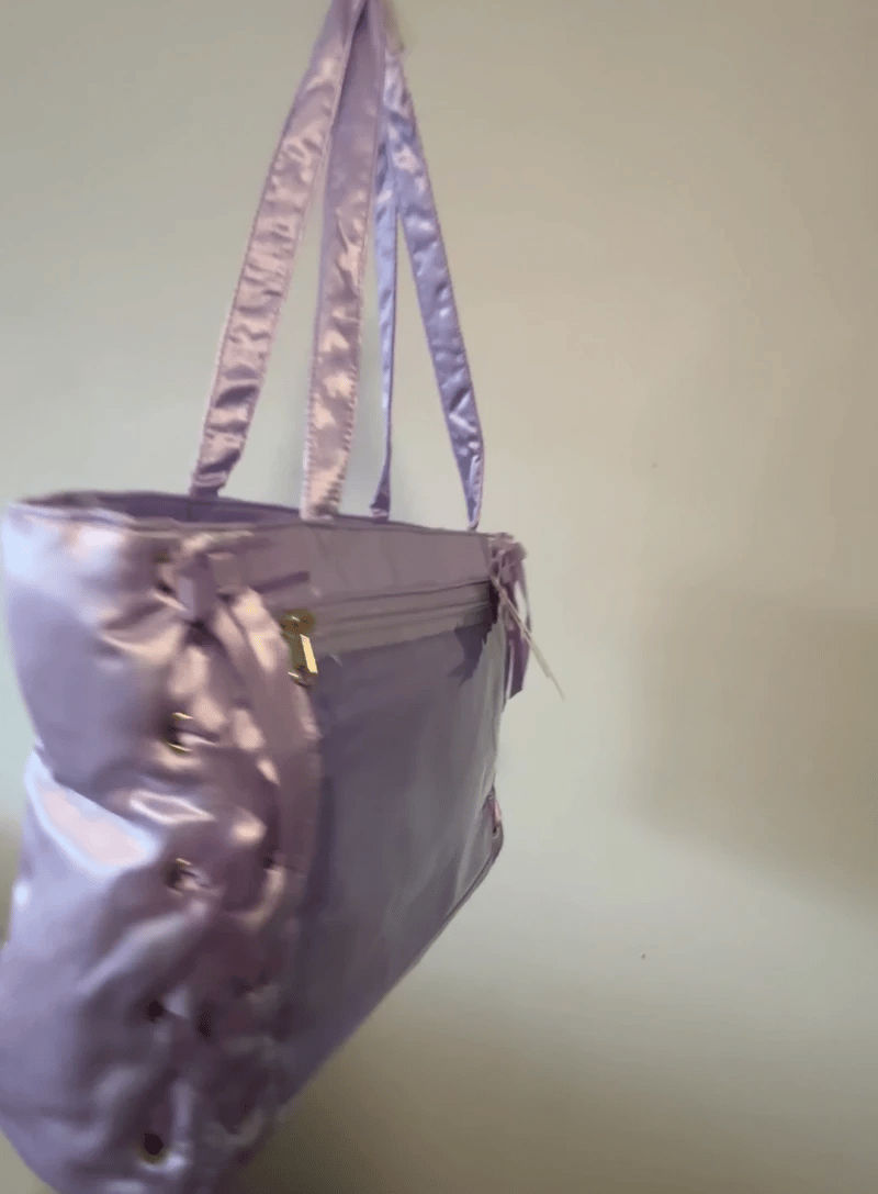 A lavender satin pin bag with side lacing and silver-tone hardware, shown hanging against a plain off-white wall.