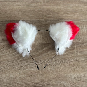 A pair of cat ears headband featuring fluffy white faux fur and bright red fabric tops, attached to a thin black wire band, shown on a light brown wood grain surface.