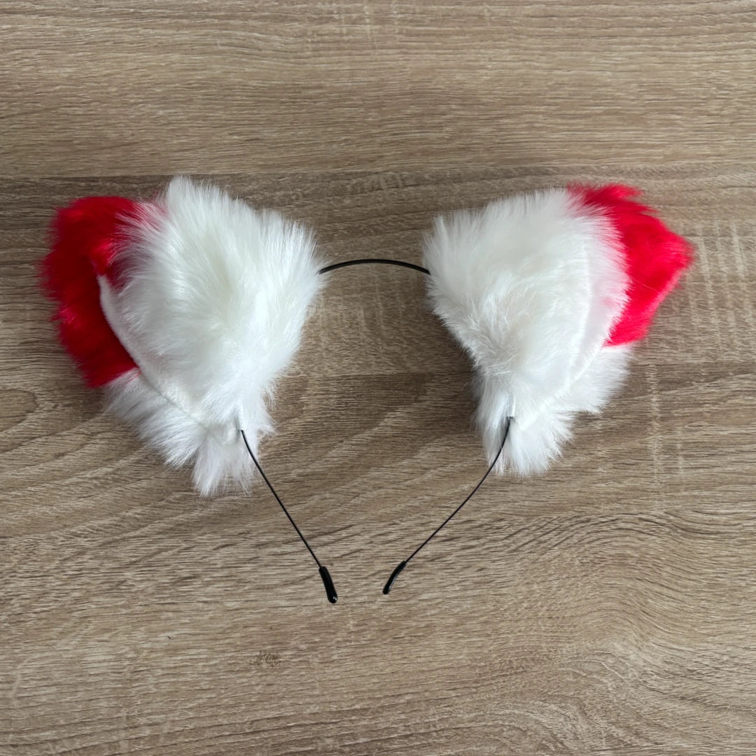 A pair of cat ears headband featuring fluffy white faux fur and bright red fabric tops, attached to a thin black wire band, shown on a light brown wood grain surface.
