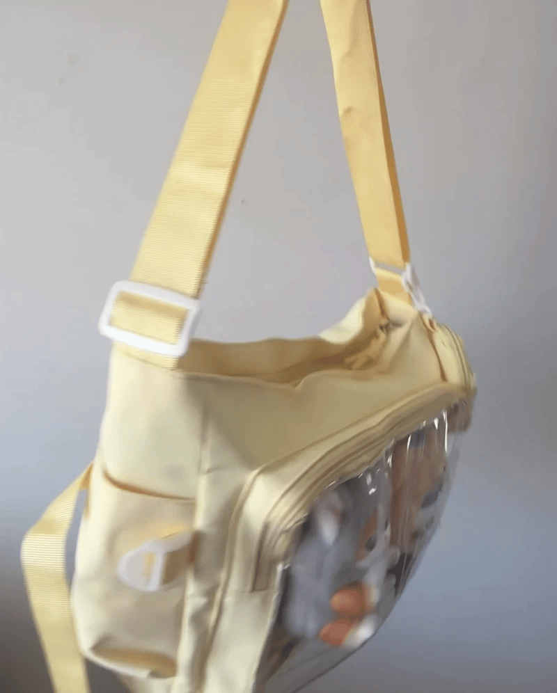 A pastel yellow Ita shoulder bag with a clear plastic front window and a matching yellow webbed strap with a white plastic buckle, shown against a plain light gray wall.
