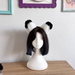 A short black wig with fluffy black-and-white cat ears is displayed on a white mannequin head, set on a wooden table against a light wall with framed pictures.