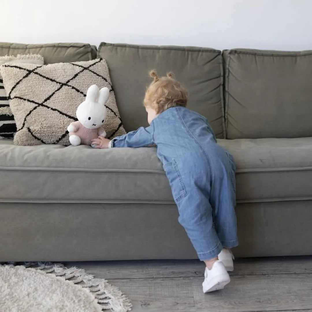 Child in blue outfit reaching for a pink plush toy on a gray sofa.