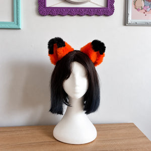 A short black bob wig with fluffy cat orange and black animal-style ears on a white mannequin head, displayed on a wooden table against a light wall with framed pictures.