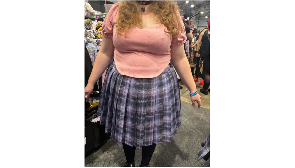 A person standing indoors wearing a pink puff-sleeve top and a blue-gray plaid pleated skirt, photographed in what appears to be a store or event space.