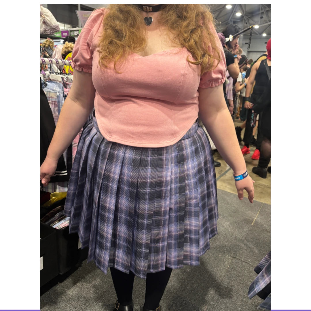 A person wearing a pink short-sleeve top and a blue-gray plaid pleated skirt, standing in a crowded indoor retail or convention space.
